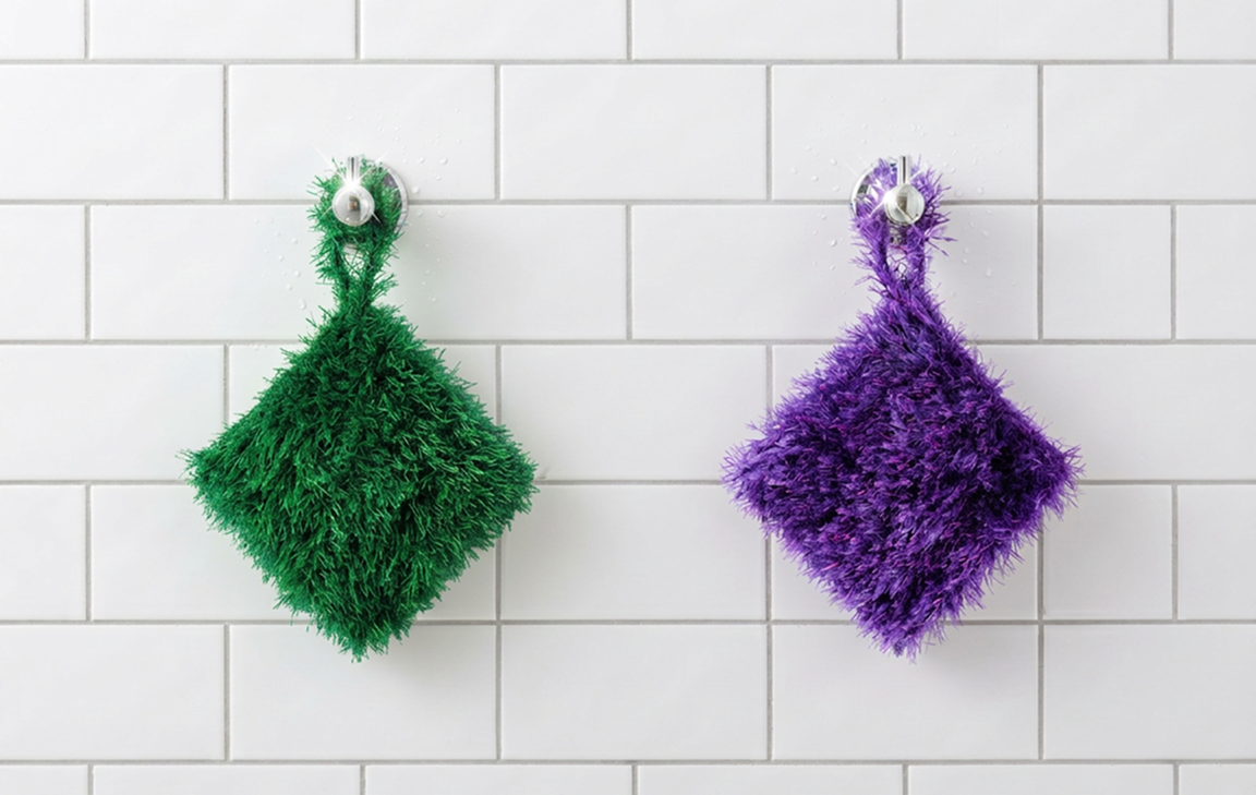 Green and purple Loofi Baby scrubbers hanging from suction hooks on white subway tile