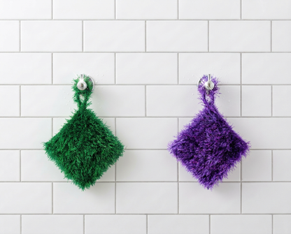 Green and purple Loofi Baby scrubbers hanging side by side on a white tiled shower wall